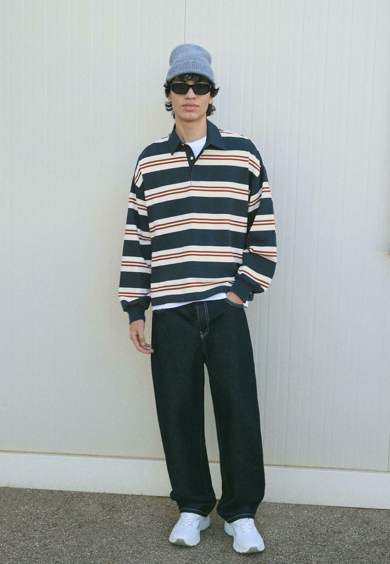 Marineblau- und weiß gestreiftes Langarm-Polo-Shirt mit roten Akzenten, kombiniert mit lockeren schwarzen Jeans und weißen Sneakers, abgerundet mit einer grauen Mütze.