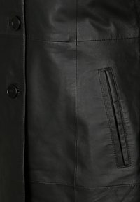 Detail einer schwarzen Lederjacke mit großen Knöpfen, horizontal genähten Panels und einer seitlichen Tasche mit glatter Textur.