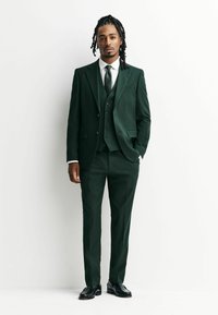 Tmavě zelený trojdílný oblek z texturované tkaniny, se klopovým límcem, vestou s knoflíky a slim-fit kalhotami. Černé boty.