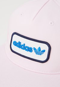 Pink cap featuring a white rectangular patch with blue embroidered "adidas" text and logo. The design includes a textured fabric.
