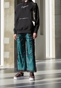 Svart hoodie med en framsida ficka och vitt logotyp, kombinerat med teal strukturerade vida byxor med svarta detaljer, samt plattformssandaletter.