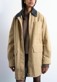Femme portant une veste beige oversize avec un col noir, un pull en maille marron et une jupe noire à motifs sur fond uni.