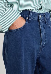 Denim kavbojke temno modre barve, z visokim pasom, petimi žepi in klasičnim krojem. V kombinaciji z zeleno-bež črtasto srajco.