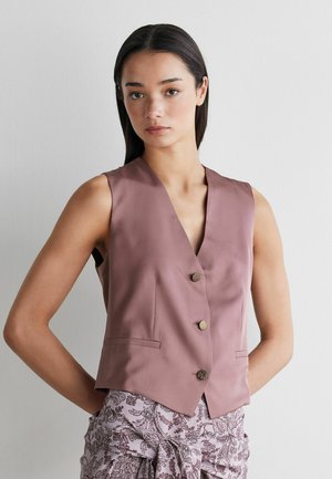 Femme aux longs cheveux noirs portant un gilet mauve sans manches boutonné et une jupe claire à motifs, debout avec les mains derrière le dos.