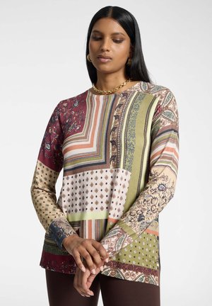 Donna con lunghi capelli neri che indossa una maglia a maniche lunghe patchwork multicolore e una collana a catena d'oro, con le mani delicatamente intrecciate davanti.