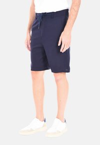 Pantaloni corti blu navy realizzati in un tessuto leggero, con una vita elasticizzata dotata di cordoncino e tasche laterali. Abbinati a sneakers bianche.
