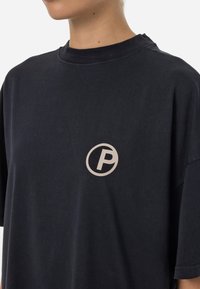 Juodas medvilninis marškinėliai su trumpomis rankovėmis, priekyje turintys apvalų logotipą su stilizuotu „P“ šviesesnėje atspalvyje. Lygus paviršius.