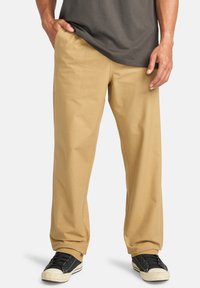 Pantalones de algodón beige con un corte holgado, que cuentan con bolsillos laterales y traseros, combinados con zapatillas negras y una camiseta gris.