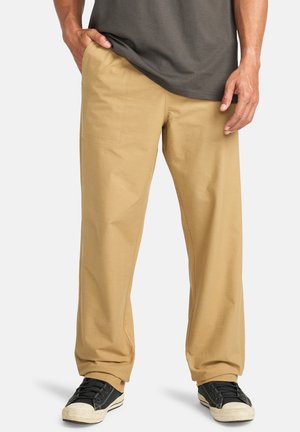 Pantaloni di cotone beige con una vestibilità ampia, dotati di tasche laterali e posteriori, abbinati a sneakers nere e una maglietta grigia.