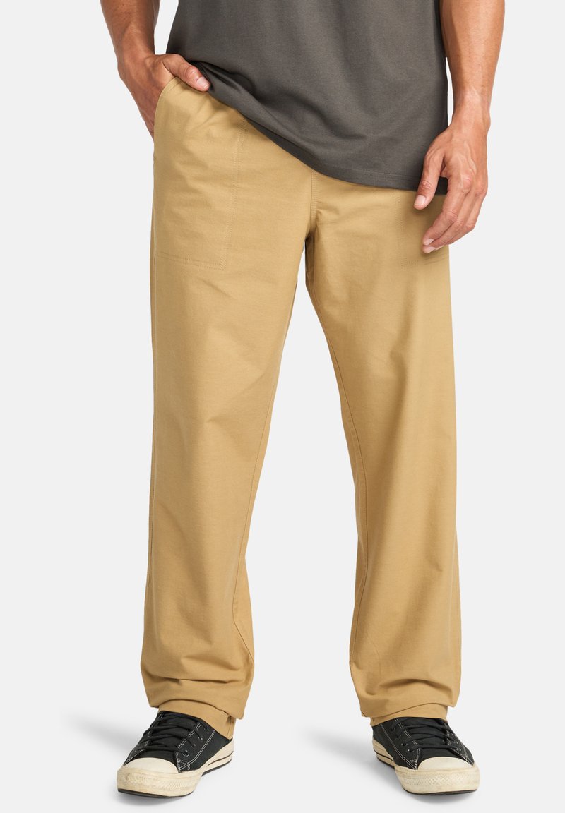 Pantalones de algodón beige con un corte holgado, que cuentan con bolsillos laterales y traseros, combinados con zapatillas negras y una camiseta gris.