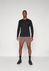 Schwarzes langärmliges Sporthemd und hellbraune Shorts, kombiniert mit schwarzen Socken und Schuhen. Merkmale sind ein figurbetontes Design und Logodetails.