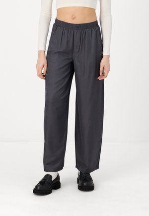 Woman wearing loose dark gray elastic-waist pants, white long-sleeve cropped top, white socks, and black loafers standing on white floor.