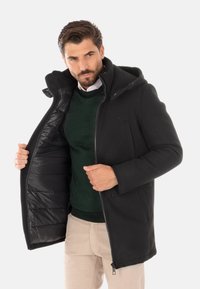 Cappotto nero con chiusura a zip e cappuccio, dotato di un interno imbottito. Indossato sopra un maglione verde e pantaloni chiari. Design strutturato e linee pulite.
