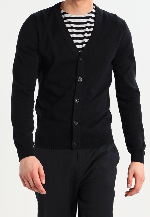 Homme portant un cardigan noir boutonné par-dessus une chemise rayée noir et blanc et un pantalon noir, debout avec les bras détendus.