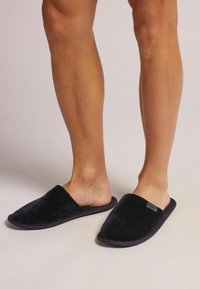 Schwarze Textilschlappen mit einer weichen Cord-Optik, einem offenen Rückendesign und kontrastierenden dunklen Gummisohlen für besseren Halt.