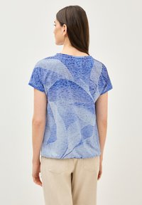 Top de manga corta azul con un diseño abstracto y fluido. Tejido suave, escote redondo y un corte relajado, combinado con pantalones beiges.