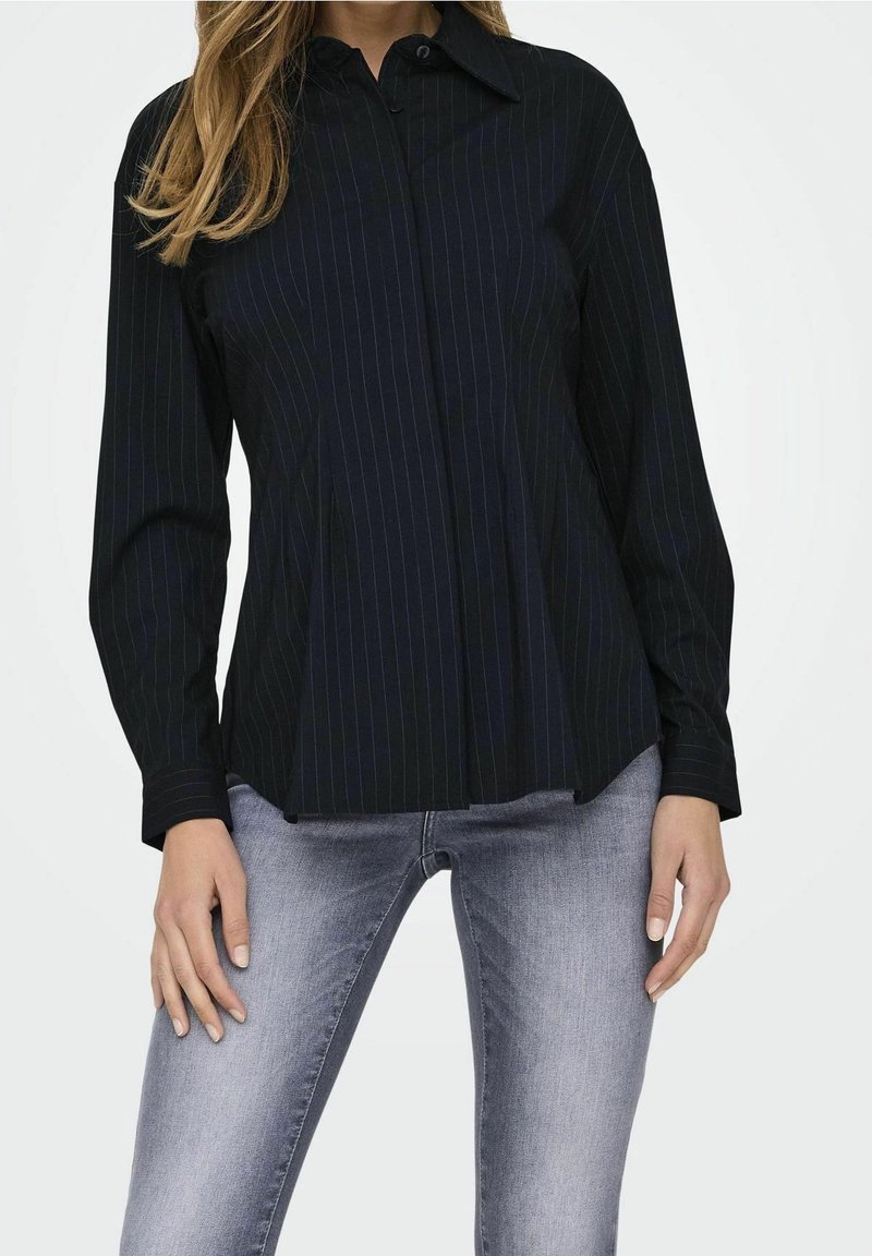 Femme portant une chemise noire à fines rayures à manches longues et un jean gris clair délavé, debout les mains détendues le long du corps.