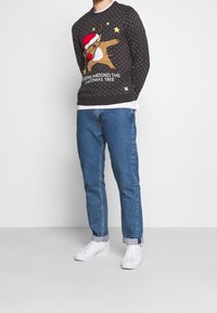 Maglione grigio scuro con motivo renna, motivo a pois, accenti bianchi, texture accogliente, abbinato a jeans blu e sneakers bianche.