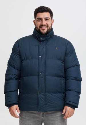 Un homme souriant porte une veste d’hiver matelassée bleu marine à col montant et un pantalon gris, sur un fond clair uni.