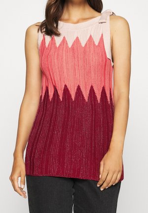 Femme portant un haut en tricot sans manches avec un motif en zigzag rose, corail et bordeaux, associé à un pantalon foncé.