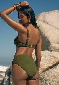 Olivno zelena bikini komplet z teksturirano tkanino, nastavljivimi trakovi in zlatim kovinskimi dodatki. Hlačke z visokim pasom nudijo popolno pokritost.