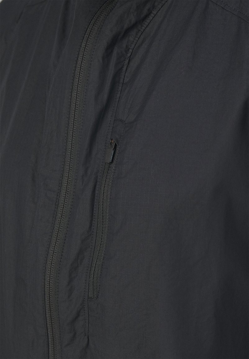 Schwarze leichte Jacke mit feiner Textur, ausgestattet mit einem zentralen Reißverschluss und einer kleinen Reißverschlusstasche auf der linken Seite.