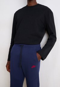 Person som bär en svart långärmad skjorta och blå Nike-svetterbyxor med dragsko, med ena handen i fickan, mot en enfärgad bakgrund.