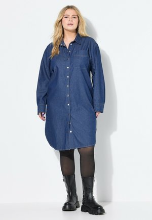 Robe chemise en denim bleu foncé avec manches longues, fermeture à boutons sur le devant, une poche poitrine et une coupe ample, associée à des bottines noires.