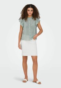 Blusa de encaje verde claro con mangas con volantes, combinada con una falda lápiz blanca. La modelo lleva sandalias planas de tejido blanco y está de pie sobre un fondo neutro.