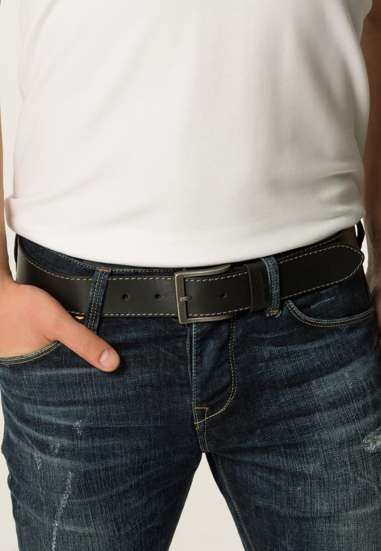 Czarny skórzany pasek z metalową klamrą w kolorze srebrnym, z detalami przeszycia i fakturowaną powierzchnią, noszony na niebieskich dżinsach.