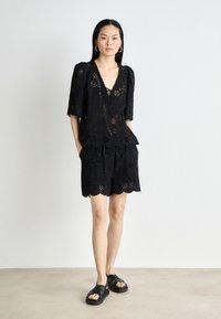 Haut en dentelle noire à œillets avec manches bouffantes, décolleté en V ouvert et ourlet festonné, associé à un short noir assorti présentant des détails similaires et une coupe décontractée.