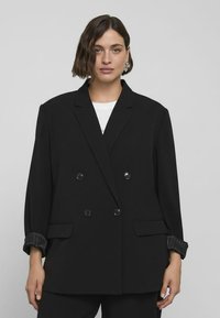 Černý dvouřadý blejzr s špičatými klopami, velkými knoflíky a texturovanými manžetami. Vyznačuje se volným střihem a dvěma předními kapsami.