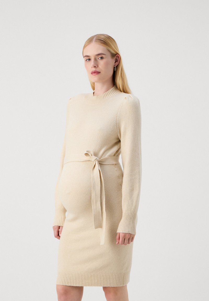 Rochie de maternitate tricotată, de culoare bej deschis, cu mâneci lungi, manșete ribbed și talie încinsă, având o textură moale și o siluetă ajustată.