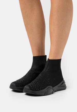 Piernas con zapatillas negras estilo calcetín con suela lisa y pequeños puntos reflectantes en el tejido de punto.