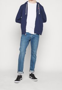 Mörkblå zip-up huvtröja med dragsnören, vit t-shirt under, ljusblå jeans och svarta Vans sneakers med vita accenter.