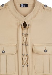 Beige jacket with lace-up front, buttoned flap pockets on both sides, epaulets with buttons on shoulders, and a collarless neckline.