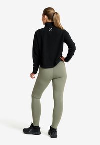 Svart fleece-pullover med hög krage, prydd med en vit logotyp. Kombinerad med olivgröna leggings och svarta träningsskor. Slät textur.