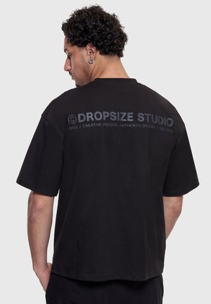 Mees lokkis juustega, kandes musta üle suurte mõõtmetega T-särki, millel on seljal kiri "DROPSIZE STUDIO", seisab tavalise heleda tausta vastu.