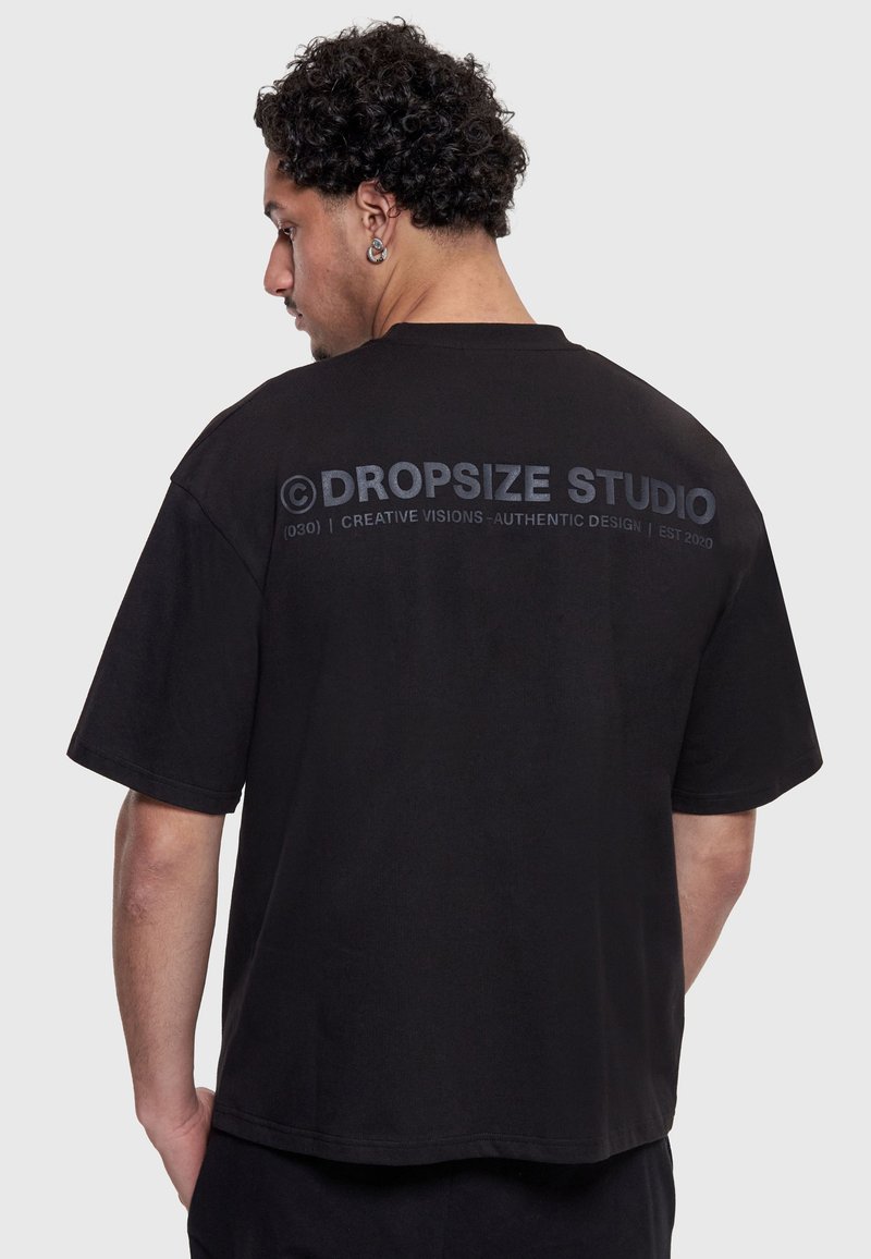 Homme aux cheveux bouclés portant un T-shirt noir oversize avec le texte « DROPSIZE STUDIO » dans le dos, debout devant un fond clair uni.