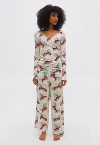 Μια γυναίκα στέκεται ξυπόλητη φορώντας μακρυμάνικη μπλούζα και φαρδιά παντελόνια σε εκρού χρώμα με μεγάλο ρόζ και πράσινο λουλουδάτο σχέδιο.