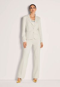Vrouw in een op maat gemaakt off-white pak met bijpassende blazer en broek, gecombineerd met gouden hoge hakken, staande voor een effen lichte achtergrond.