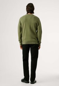 Olivinės spalvos tekstūruotas megztinis su gilia apykakle, dėvimas ant juodų kelnių ir juodų batų, matomas iš nugaros, prieš šviesų foną.