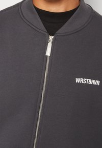 Šedá bunda na zip s žebrovanou strukturou, zaobleným límcem a bílým nápisem "WRSTBHV". Kovový zip s elegantním designem.