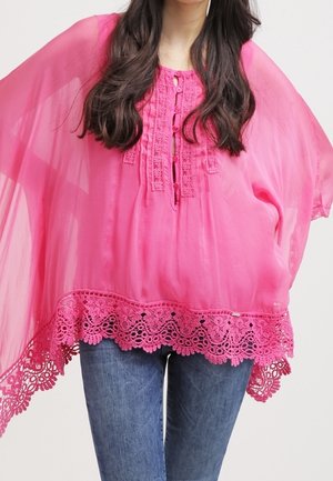 Femme portant un chemisier rose transparent à bordure en dentelle et boutons, associé à un jean bleu, montrant seulement le torse et le haut des jambes.