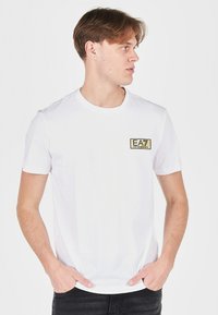 Bílá bavlněná trička s krátkými rukávy, kulatým výstřihem a zlatou vyšívanou logem EA7 na levé straně hrudi. Jednoduchý design, bez vzorů.