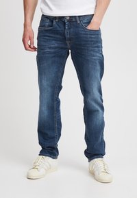Blå denimjeans med rette ben, med lommer foran og bak, sømmer med stikninger og en falmet vasketekstur. Kombinert med hvite joggesko.