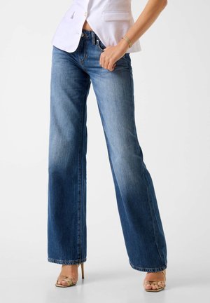 Femme portant un jean bleu taille moyenne à jambes larges, un blazer blanc, un bracelet en or et des sandales à talons hauts dorées à brides, debout devant un fond blanc.