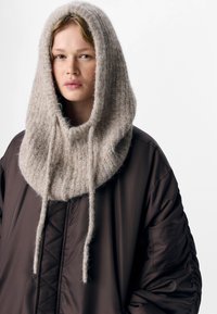 Beige knitted hood with a soft texture, framing the face, attached to a dark brown puffy jacket with textured fabric and visible seams.
