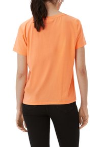 Orange Baumwoll-T-Shirt mit kurzen Ärmeln, lockerer Passform, rundem Ausschnitt und sichtbarem Nähen. Getragen mit schwarzen Hosen, von hinten betrachtet.