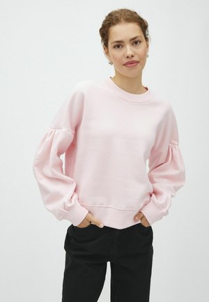Jeune femme aux cheveux clairs et bouclés porte un sweat-shirt rose clair avec des manches froncées et un pantalon noir, debout les mains dans les poches.